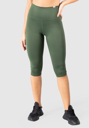 Leggings capri olive avec une taille haute, en tissu extensible et lisse. Portés avec des chaussures de sport noires.