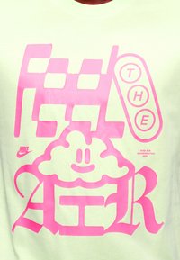 Φωτεινό πράσινο μπλουζάκι με μεγάλο ροζ κείμενο γραφικών που γράφει "FEED THE AG" και ένα χαμογελαστό σύννεφο. Λογότυπο Nike στην επάνω αριστερή γωνία.