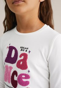 Fehér, hosszú ujjú ing, textúrált, színes betűkkel, melyek a "DANCE" szót írják. Tartalmazza a "DREAM BIG &" feliratot fekete szöveggel felette.