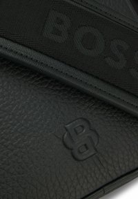 Borsa in pelle nera testurizzata con tracolla intrecciata con logo "BOSS". Include un dettaglio con logo in rilievo. Design compatto e angolare.