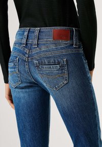 Tumši zili džinsa bikses ar pieguļošu dizainu, raksturīgas ar teksturētu apdari, aizmugures kabatām un brūnu ādas uzlīmi pie jostasvietas.