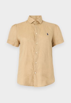 Γυναίκα που φοράει μπεζ πουκάμισο με κοντό μανίκι και κουμπιά, άσπρες κοντές βερμούδες, άσπρα σανδάλια, κρατώντας μια μικρή λευκή τσάντα, στέκεται μπροστά σε ένα μονόχρωμο φόντο.