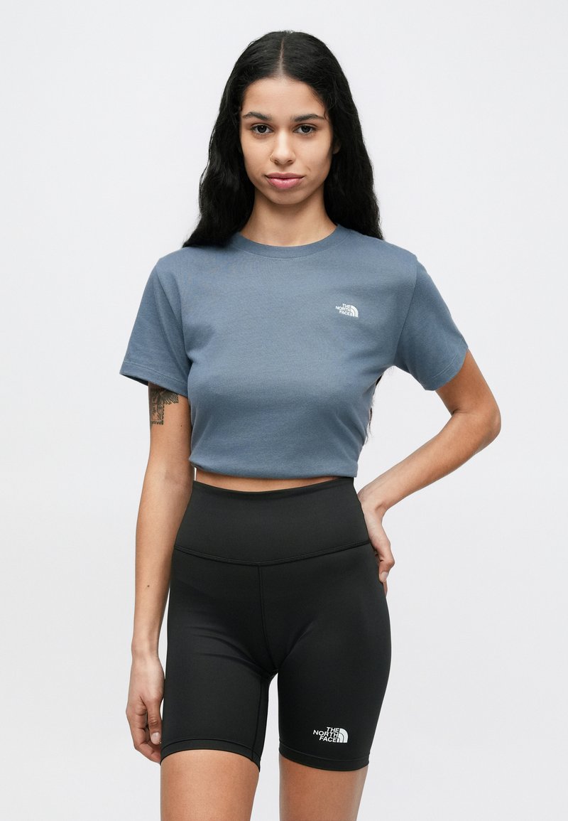 Kvinde med langt mørkt hår iført en blå crop top og sorte højttaljede cykelshorts, begge med The North Face-logoer.