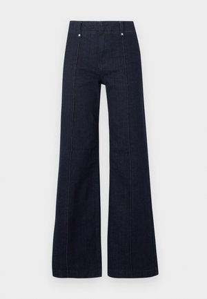 Jean bleu foncé taille haute à jambes larges avec coutures devant et passants pour ceinture sur un fond clair uni.