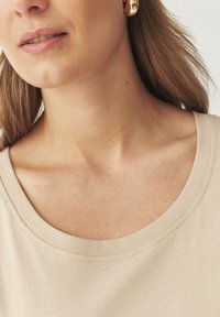 Haut beige avec un col large et arrondi. Le tissu semble doux et a une texture lisse. Comprend des détails de couture visibles.