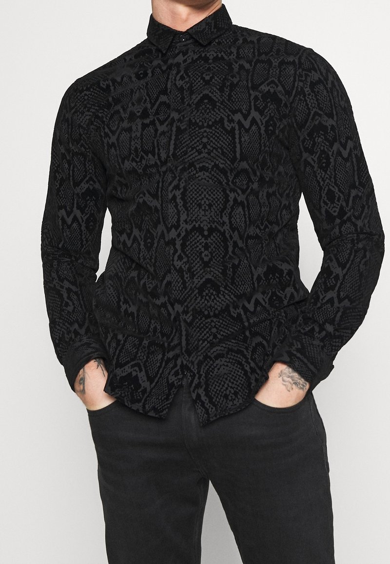 Homme portant une chemise noire à manches longues avec un motif texturé de serpent et un pantalon noir, les mains partiellement dans les poches, des tatouages visibles sur les poignets.