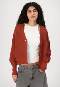 Kort rustfarget cardigan med teksturert strikkedesign, med cropped lengde, nedre skuldre og fire runde knapper foran.
