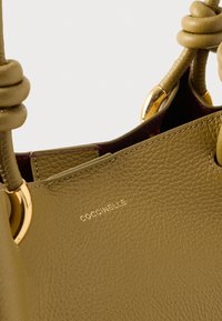 Torba od kožne kože smeđe boje s čvorastim ručkama, zlatnim metalnim detaljima i utisnutim imenom marke "Coccinelle" na prednjoj strani.