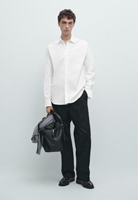 Camicia bianca con bottoni, pantaloni neri e borsa nera con maglione grigio. Il modello è in piedi su uno sfondo chiaro con una postura rilassata.