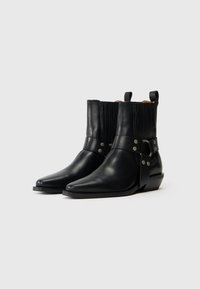 Botas de cuero negras hasta el tobillo con punta afilada, que presentan una parte superior acanalada, una correa ancha con acentos metálicos y un tacón bajo apilado.