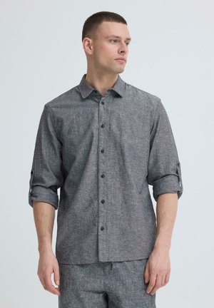 Mand iført langærmet grå skjorte med knapper, ærmerne rullet op til albuerne, stående mod en ensfarvet hvid baggrund, kiggende mod højre.