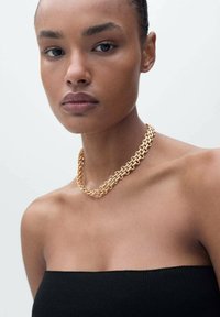 Collana in oro con anelli intrecciati, indossata su pelle nuda, sopra un top senza spalline nero. Texture liscia, finitura lucida.