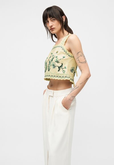 Νεαρή γυναίκα με τατουάζ που φοράει ένα χρωματιστό κέντημα σε ανοιχτό κίτρινο κοντό τοπ και ψηλόμεσα λευκά παντελόνια, στέκεται με τα χέρια στις τσέπες.
