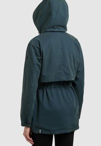 Personne portant une veste à capuche vert foncé avec une taille élastique et des manches longues, vue de dos sur un fond uni.