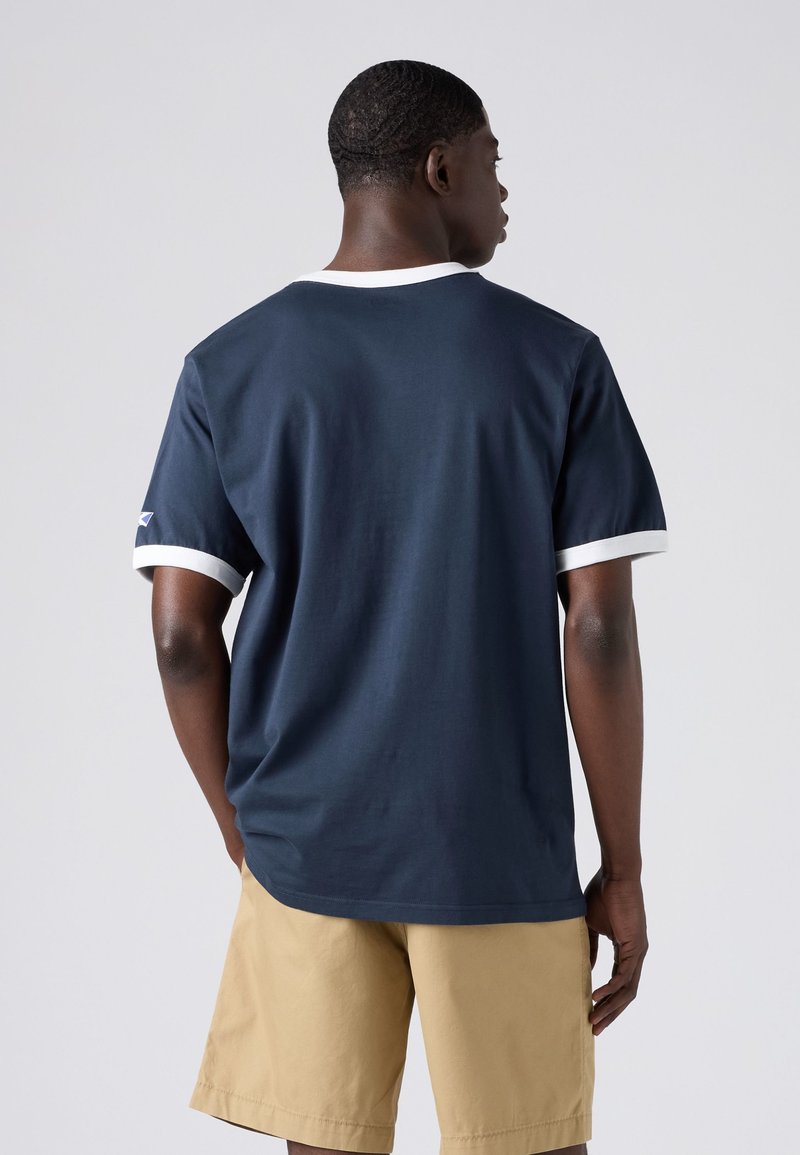 Man wearing navy blue T-shirt with white trim and beige shorts, standing facing away against a plain light background.
