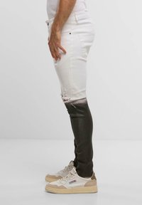 Muž v slim-fit dip-dye džínách přecházejících z bílé do černé barvy s obnošenými detaily a béžovými teniskami na neutrálním pozadí.