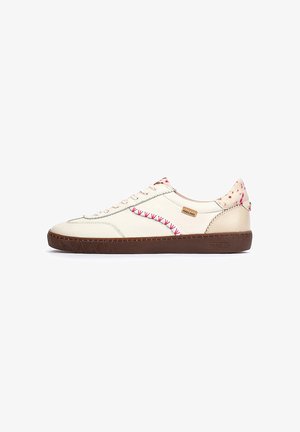 Zapatilla deportiva de color crema con suela de goma marrón, que presenta costuras rojas decorativas y un acento trasero con patrón.