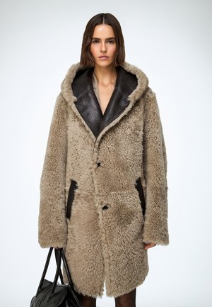 Manteau en fourrure beige avec un col châle en cuir marron, fermeture croisée à double boutonnage et poches latérales, porté avec un sac à main noir.