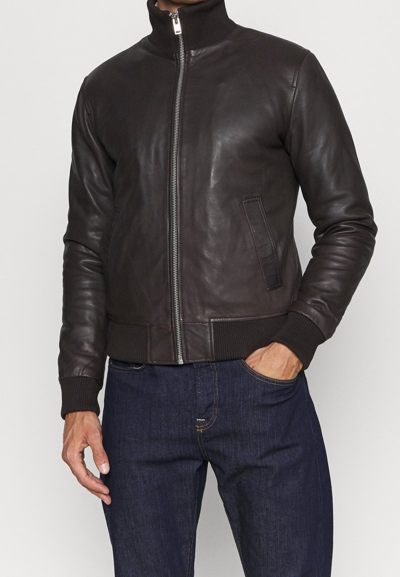 Veste aviateur en cuir marron avec un col et des poignets côtelés, dotée d'une fermeture éclair sur le devant et de poches latérales. Texture lisse, style ajusté.