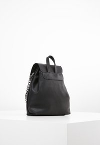 Czarny skórzany plecak z klapą przewijaną na górze, fakturowaną powierzchnią i ozdobą w formie łańcucha. Posiada górny uchwyt i pojedynczy pasek na ramię.