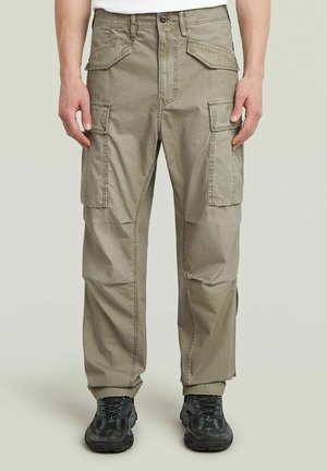 Mann trägt hellbraune Cargohosen mit mehreren Taschen und schwarzen Sneakers, steht vor einem einfarbigen hellen Hintergrund.