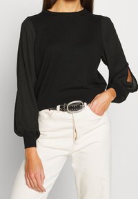 Svart tröja med figursydd nederdel och voluminösa genomskinliga ärmar, med utskärningar vid manschetterna. Bärs med vita jeans och ett spikigt bälte.