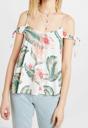 Kobieta ubrana w biały top z odkrytymi ramionami z zielono-różowym tropikalnym kwiatowym wzorem oraz jasnoniebieskie dżinsy, ozdobiona warstwowymi złotymi naszyjnikami.