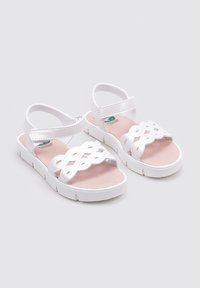 Sandalias de plástico blancas con un diseño superior entrecruzado. Cuentan con una plantilla acolchada rosa y una correa ajustable en el tobillo. Suela plana.
