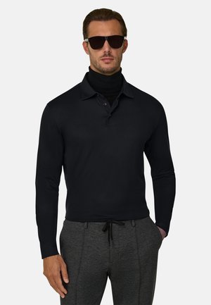 Hombre con gafas de sol negras, polo de manga larga negra sobre un jersey de cuello alto negro y pantalones gris oscuro con cordón, sobre un fondo liso.