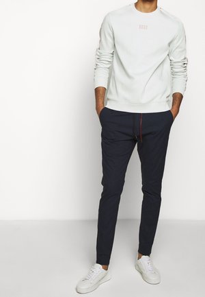 Hellgrauer Pullover mit gerippten Bündchen und kleinem Logo. Dunkelblaue Jogginghose mit rotem Kordelzug und zulaufenden Beinen. Weiße Sneaker.