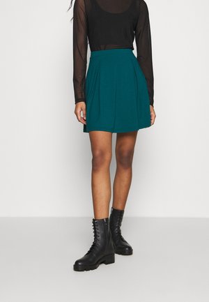 Mujer con falda tableada color verde azulado, blusa negra de manga larga y transparente, y botines negros con cordones, de pie frente a un fondo liso.