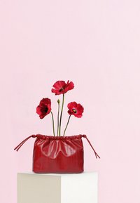 Κόκκινη τσάντα από δέρμα με κλείσιμο κορδονιού, που περιέχει τρία ζωντανά κόκκινα λουλούδια. Ομαλή υφή, τοποθετημένη σε ένα φωτεινό βάθρο πάνω σε ροζ φόντο.