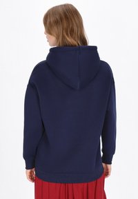 Tengerészkék kapucnis pulóver nagy kapucnival, hosszú ujjakkal és bordázott mandzsettákkal, vörös pliszírozott szoknyával viselve. Varrat nélküli hátoldali kialakítás.