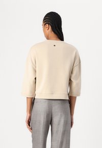 Felpa cropped di un leggero beige con maniche a tre quarti; texture morbida e design minimalista; presenta un piccolo dettaglio del logo sul retro.