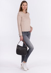 Maglia beige a maniche lunghe, jeans grigi slim, sneakers bianche, tiene una borsa nera con motivo e tracolla in pelle marrone. Sfondo semplice.