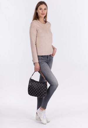 Beige langmouwen top, slanke grijze jeans, witte sneakers, houdt een zwarte handtas met patroon vast met een bruine leren schouderband. Eenvoudige achtergrond.