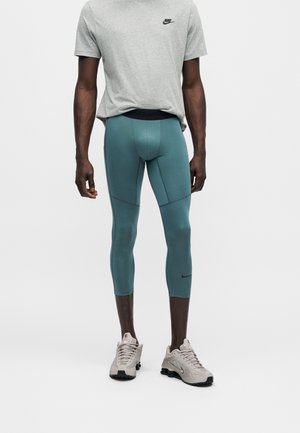 Muškarac nosi sivu Nike majicu, tirkizne kompresijske tajice skraćene iznad gležnjeva i svijetlo bež Nike tenisice, stoji na bijeloj pozadini.