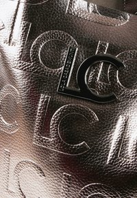 Zbliżenie na ciemną, metaliczną skórę z teksturą, z wytłoczonym wzorem "LC" oraz czarną metalową tabliczką z logo "LC" przymocowaną.