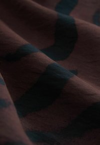 Bavlněná tkanina s hladkým povrchem, která má tmavě modrý abstraktní vzor na bohatém hnědém pozadí. Jemné záhyby zvýrazňují design.