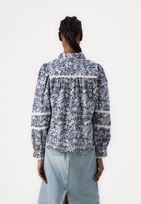 Marine-mønstret skjorte med hvite blomsterdesign, sammenbundet bak, og puffede ermer, kombinert med et lyst denim-skjørt.