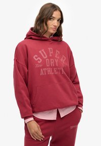 Bordeaux oversized hoodie met een voortasje, met de tekst "SUPER DRY ATHLETIC" in lichtere letters. Katoenmengsel, zachte structuur.
