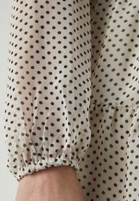 Διαφανής μπλούζα με μακριά μανίκια και μαύρες βούλες σε ανοιχτό μπεζ φόντο, με ελαστικούς στραγγαλιστές και ρομαντική σχεδίαση.