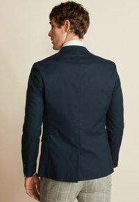 Marineblå skræddersyet blazer i glat stof, med en enkelt slids bagpå og slanke revers, parret med grå ternede bukser.