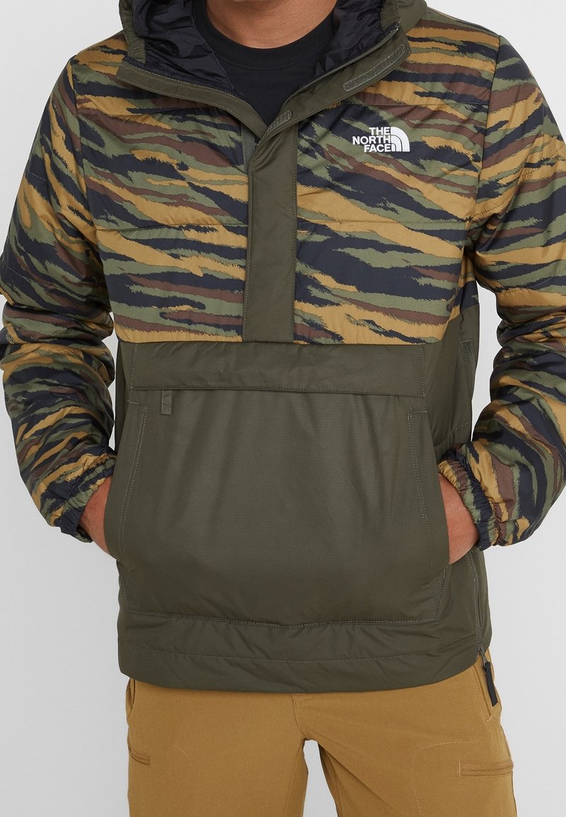 Homme portant une veste à enfiler The North Face verte et camouflage avec poche kangourou à l'avant et pantalon beige.