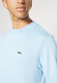 Sweatshirt en coton bleu clair avec un col rond côtelé et un petit logo crocodile brodé en vert sur la poitrine. Texture lisse.