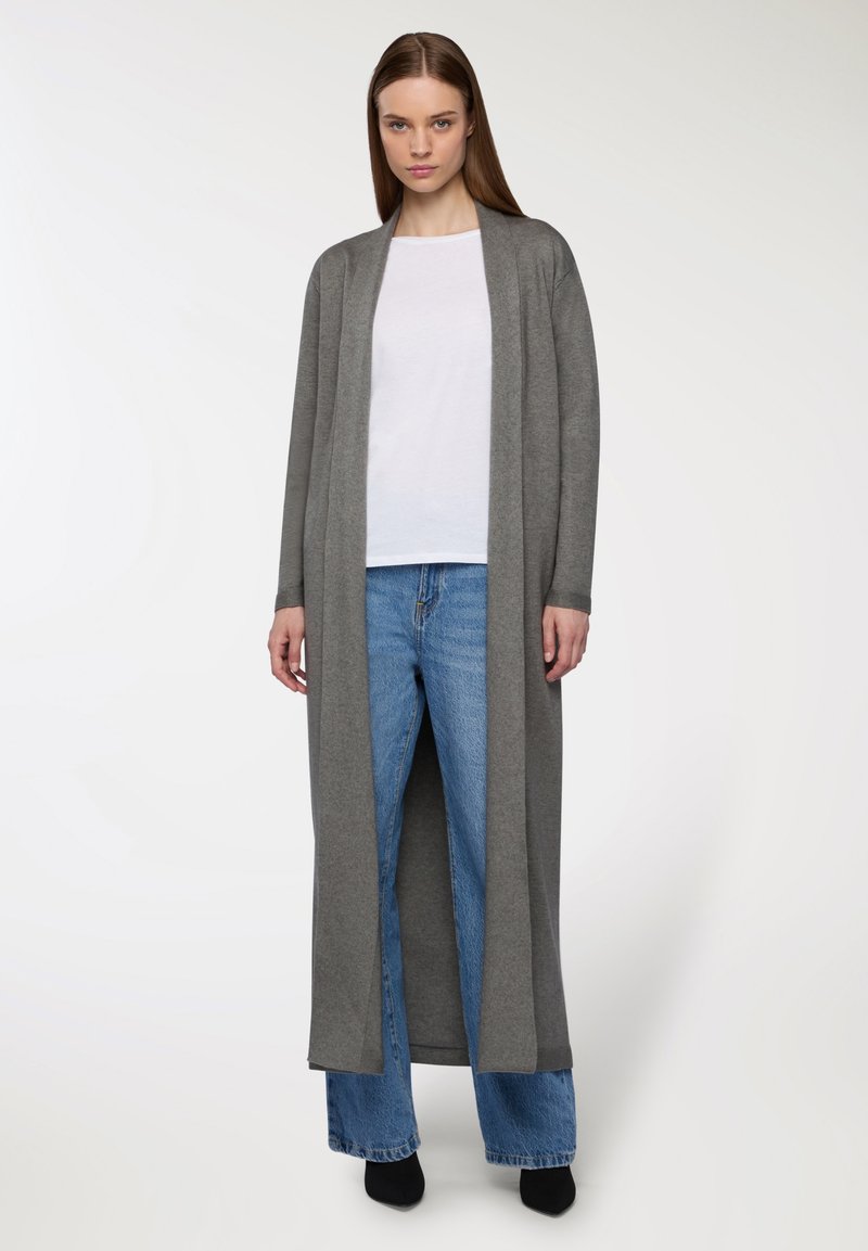 Długi kardigan z otwartym przodem w miękkim szarym materiale, z długimi rękawami, prostym krojem i bocznymi rozcięciami, połączony z białą bluzką i niebieskimi dżinsami.