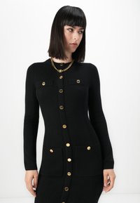 Una donna con capelli neri e lisci indossa un abito nero aderente con bottoni dorati e tasche a patta, abbinato a una collana con catena dorata voluminosa.