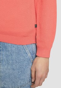 Coralfarbenes, strukturiertes Strickpullover mit geripptem Saum und Bündchen, kombiniert mit hellblauer Jeans. Kleines Markenschild auf dem Pullover sichtbar.