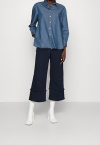 Lichtblauw denim overhemd met een borstzak, gecombineerd met marineblauwe wijde broek met franjerand en witte laarzen met hak.