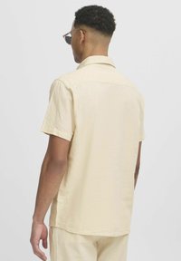 Chemise à manches courtes, de couleur beige clair, en tissu texturé. Conçue avec un col pointu et des détails au niveau du dos.